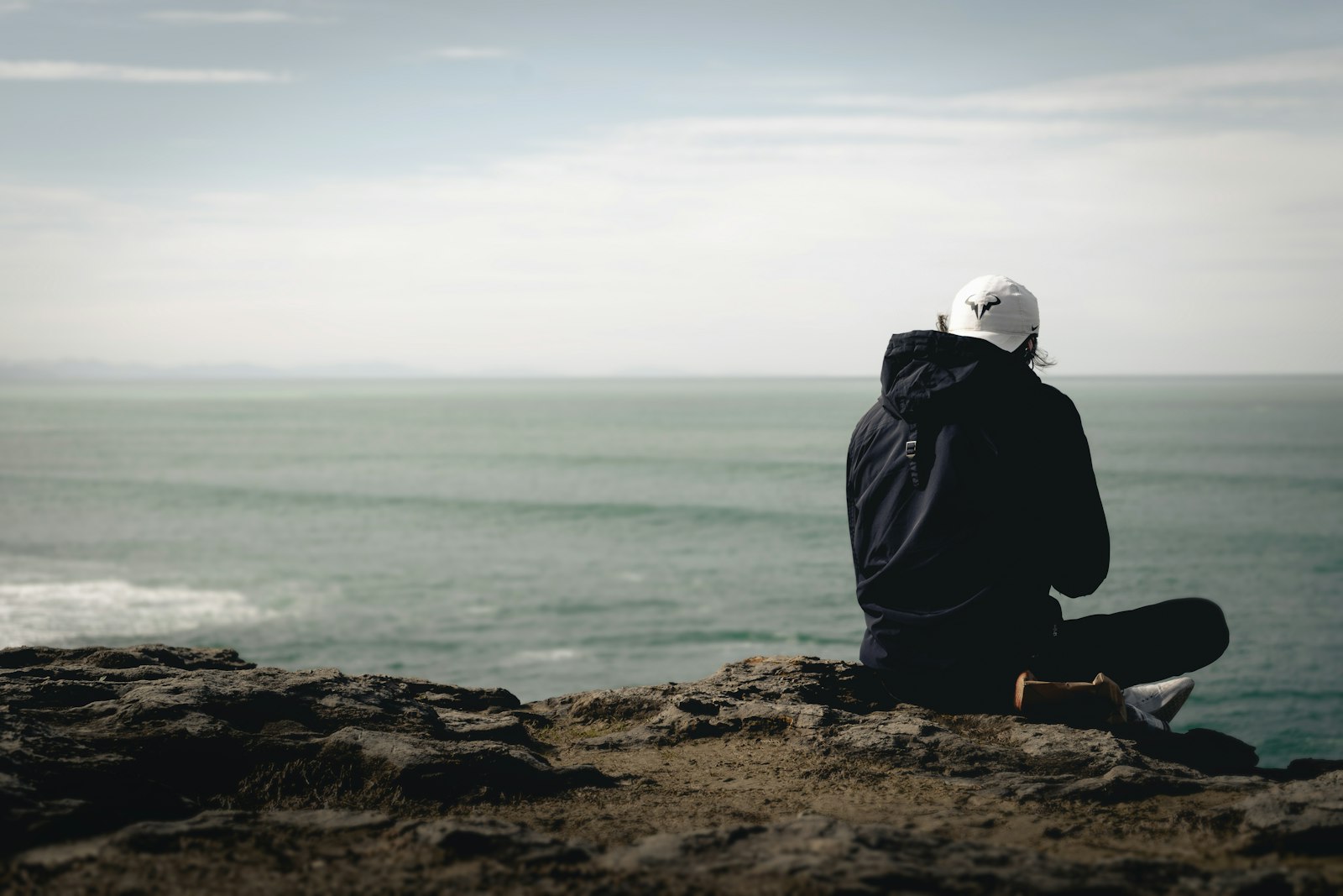 One person sitting alone on dark rocks at the shoreline, looking out at a flat grey sea.