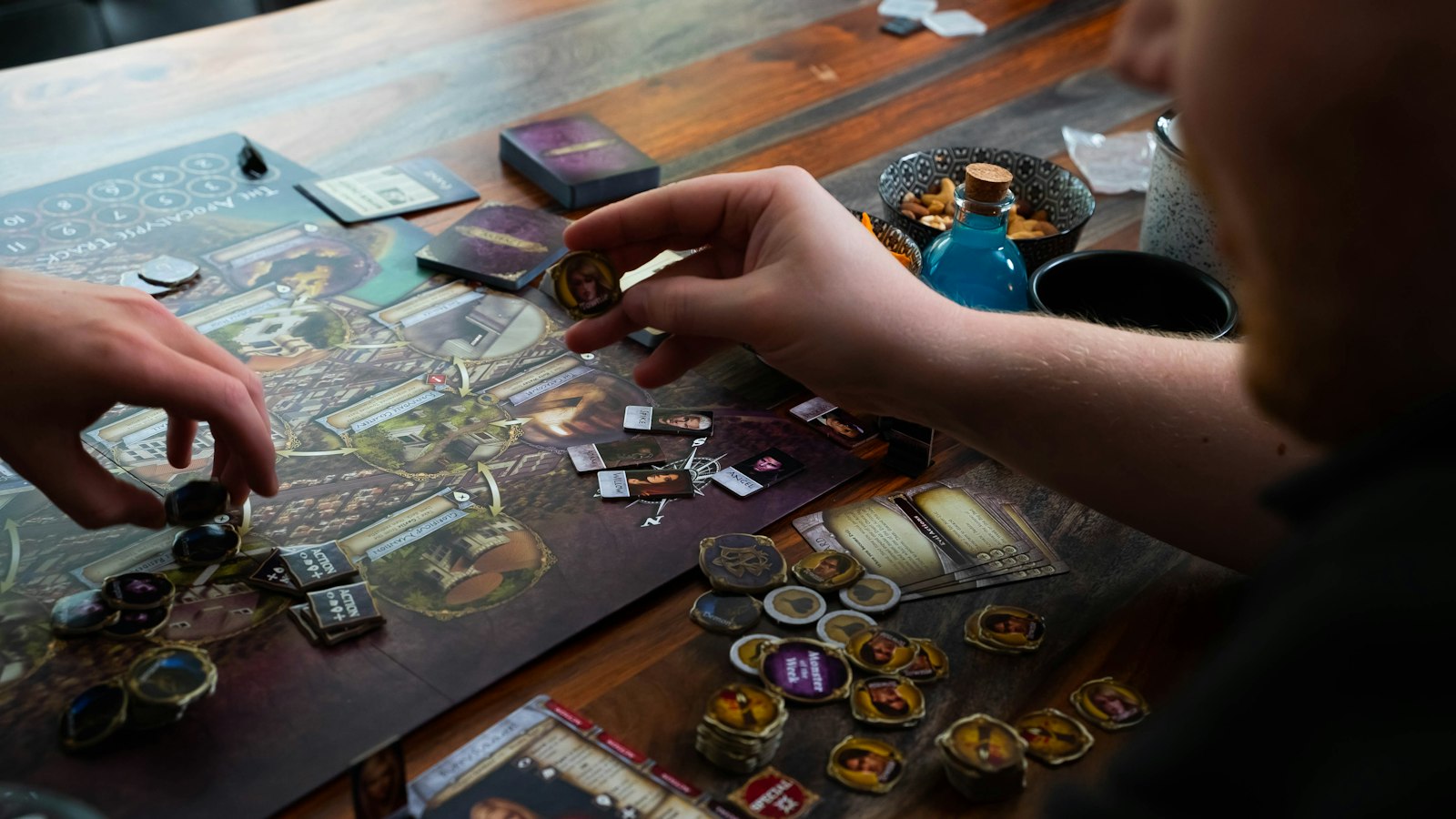 Two people's hands reaching over a board game spread across a wooden table, cards and wooden pieces between them.