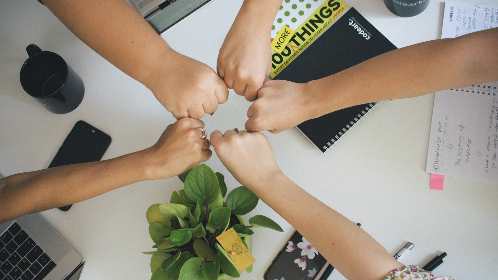 People bringing their fists together above notebooks and a laptop on a table.