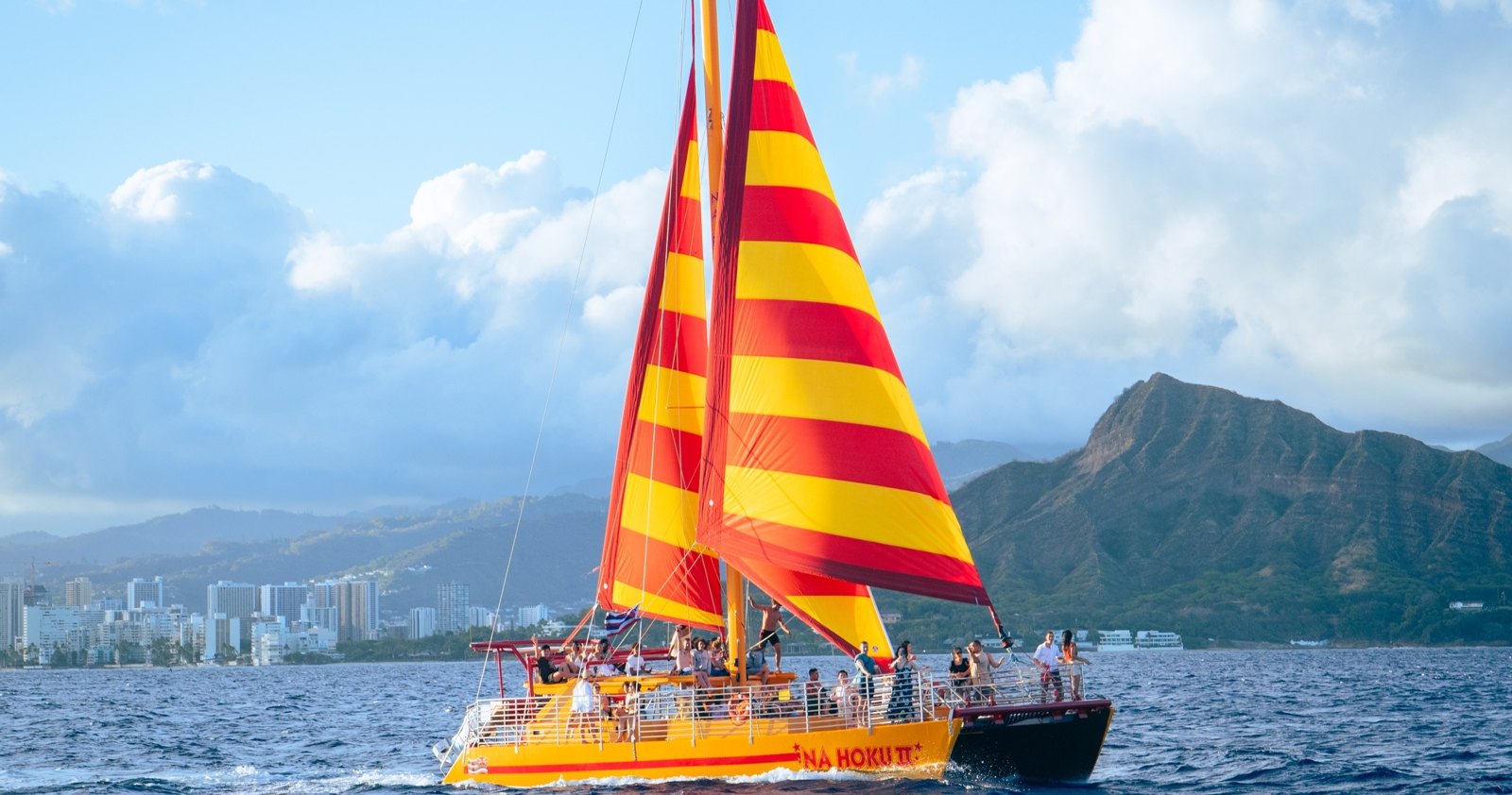 Na Hoku 2 sailing off Honolulu with red and yellow sails and Diamond Head in the background.
