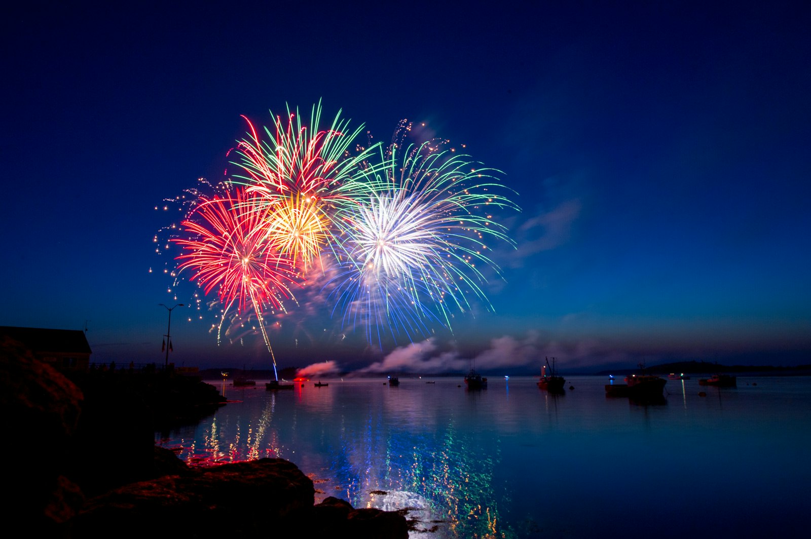 Fireworks bursting over a calm bay at dusk, with small boats moored below and the colours reflected in the water.