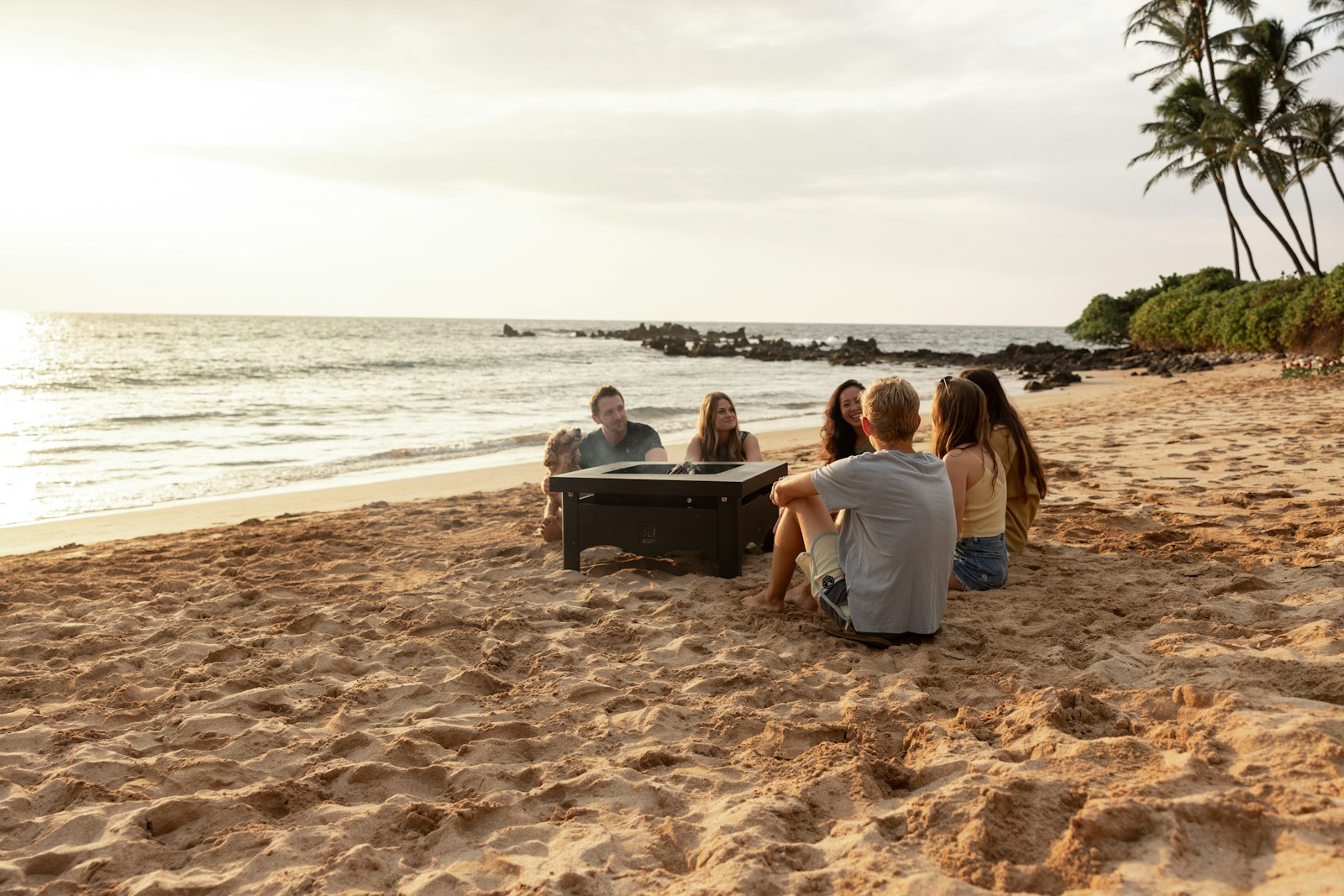 A group of friends sitting around a fire pit on a Hawaii beach at sunset, the kind of real plan people open on YKnot.