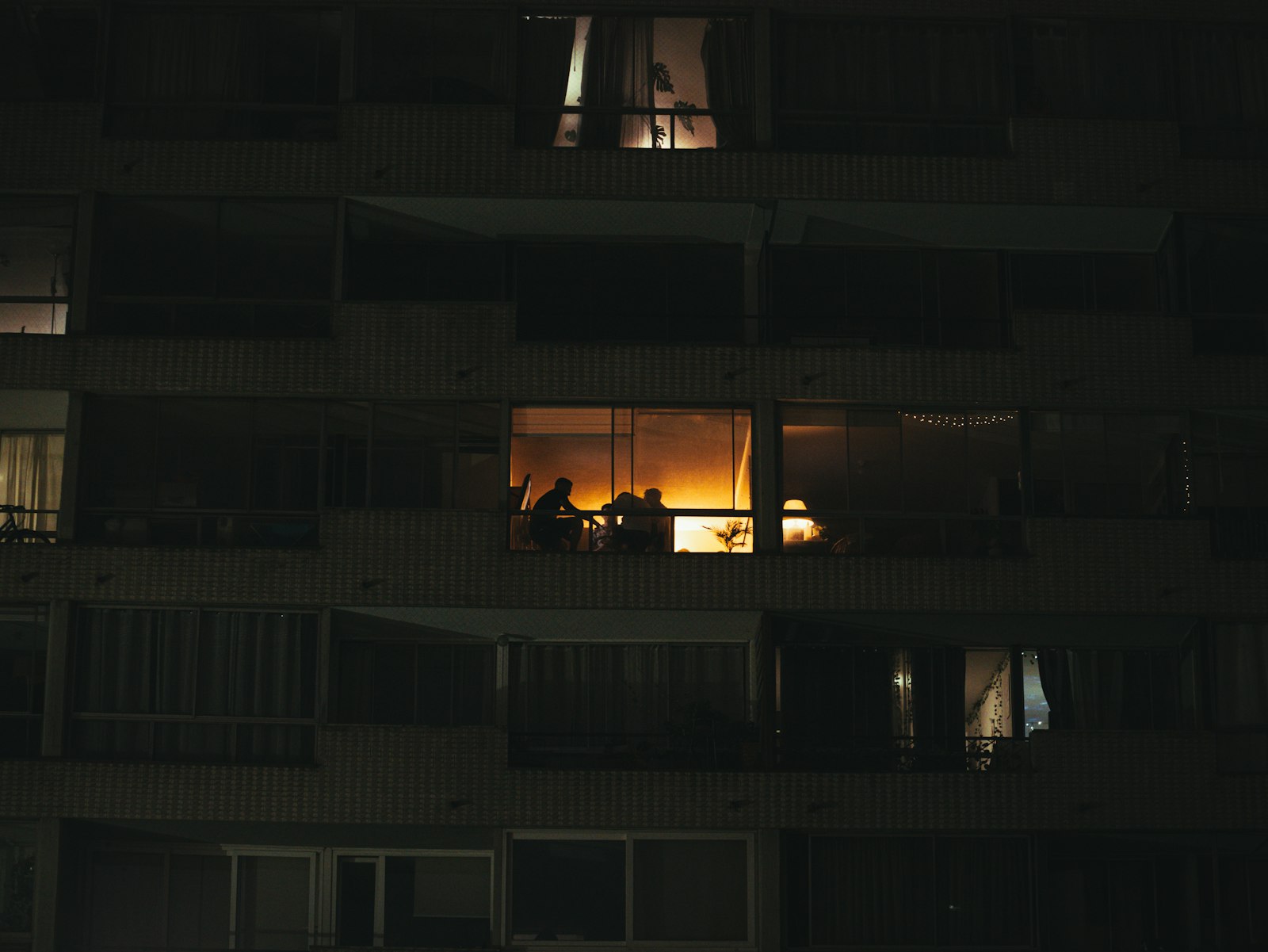 One lit apartment window at night, people gathered inside, in an otherwise dark block of flats