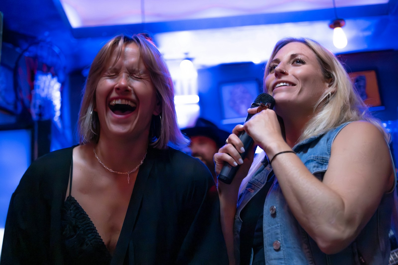A woman laughing while singing into a microphone under colored lights, karaoke in Waikiki with friends.