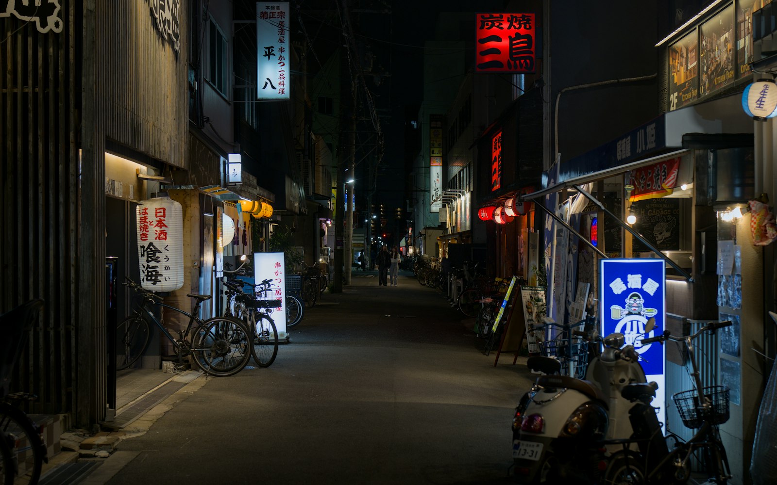 A narrow alley of small restaurants at night, lanterns and signs lit along both sides.