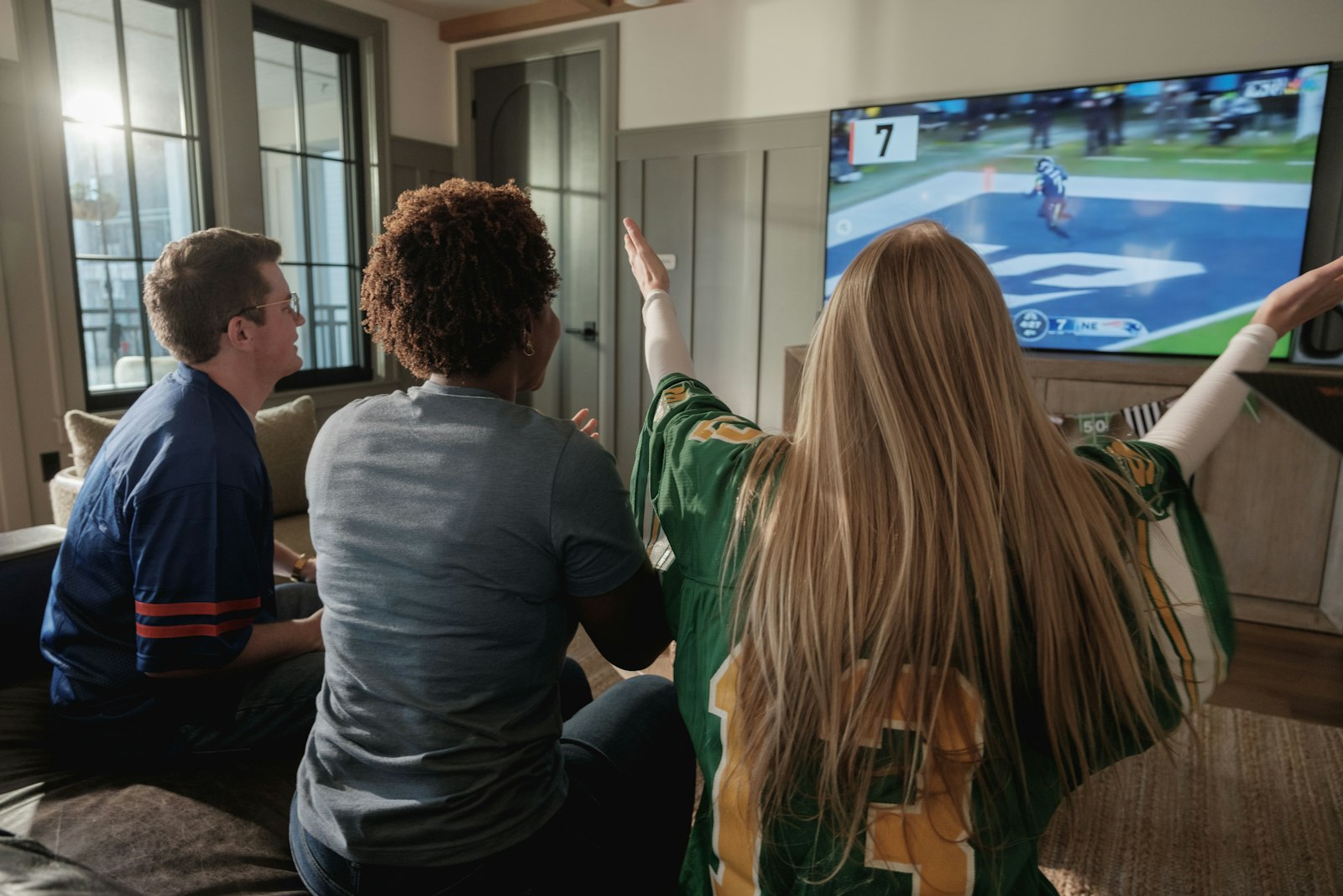 Three people on a sofa watching a baseball game on a television, one of them reacting to the play.