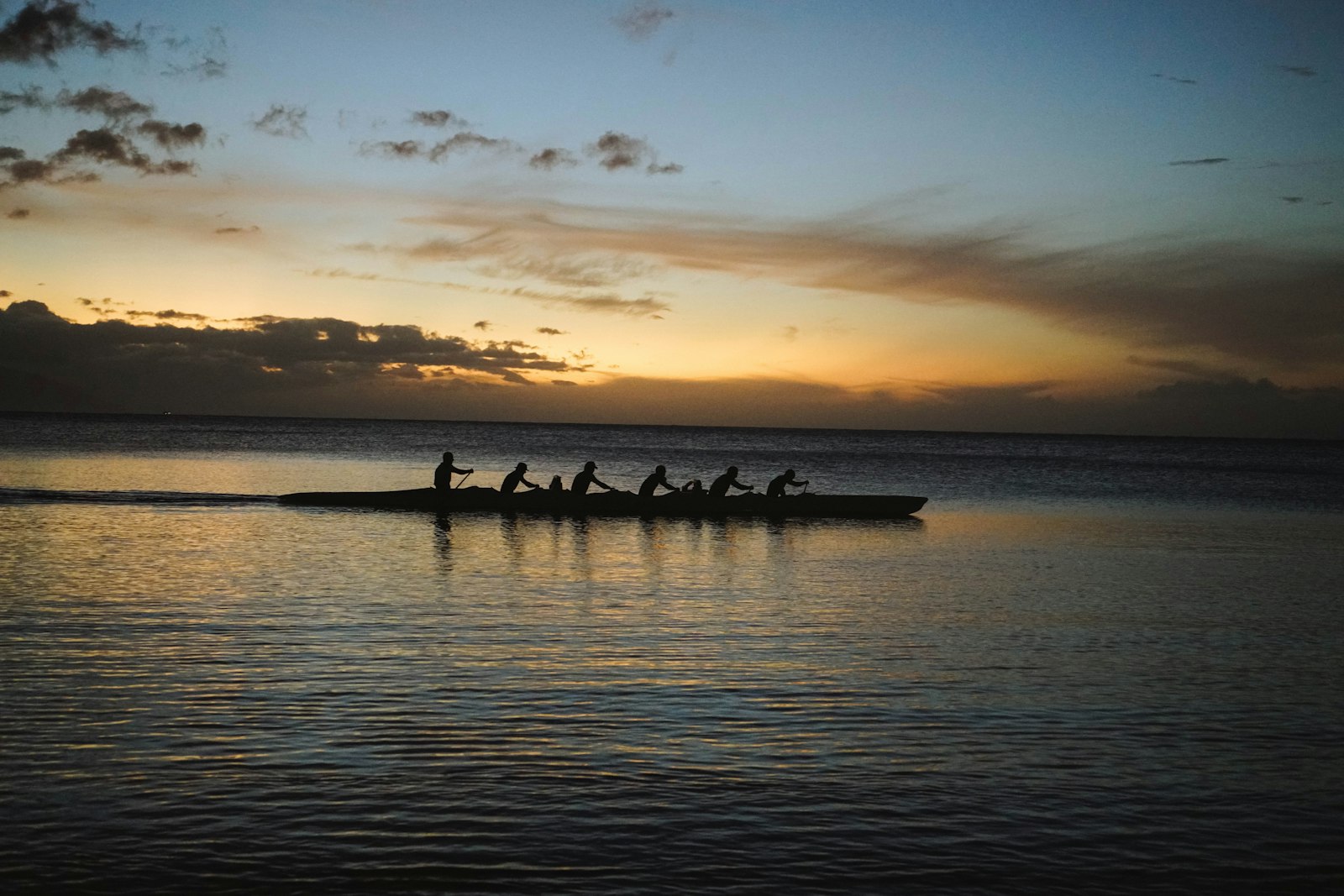 An outrigger canoe crew paddling in silhouette across flat water at sunset.