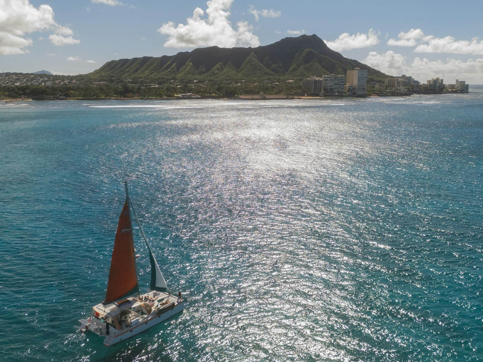 A catamaran under a red sail on the water off Waikiki, with Diamond Head and the shoreline behind it