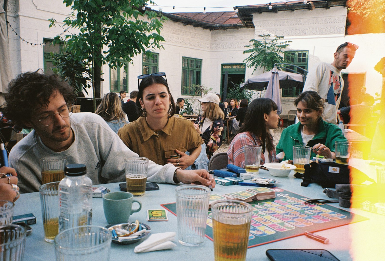 Eight or nine people around one long table mid-game, drinks and boxes pushed to the edges, several conversations going at once.