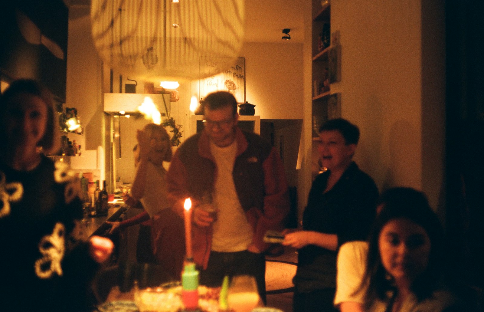 A crowded table of friends eating and talking by candlelight in somebody's front room.