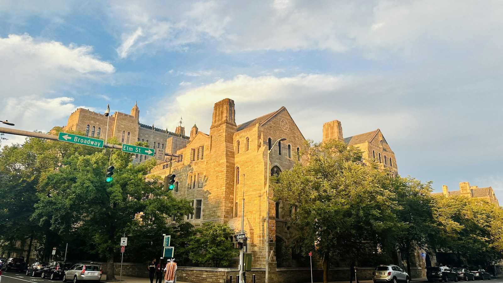 Sunlit stone buildings along a tree-lined city street, with pedestrians at the corner.