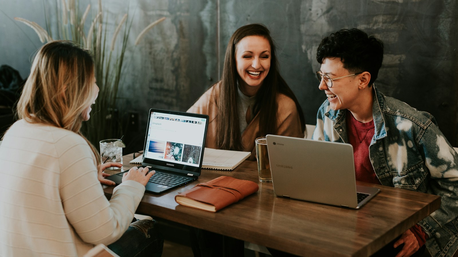 Three people talking around laptops at a wooden table.