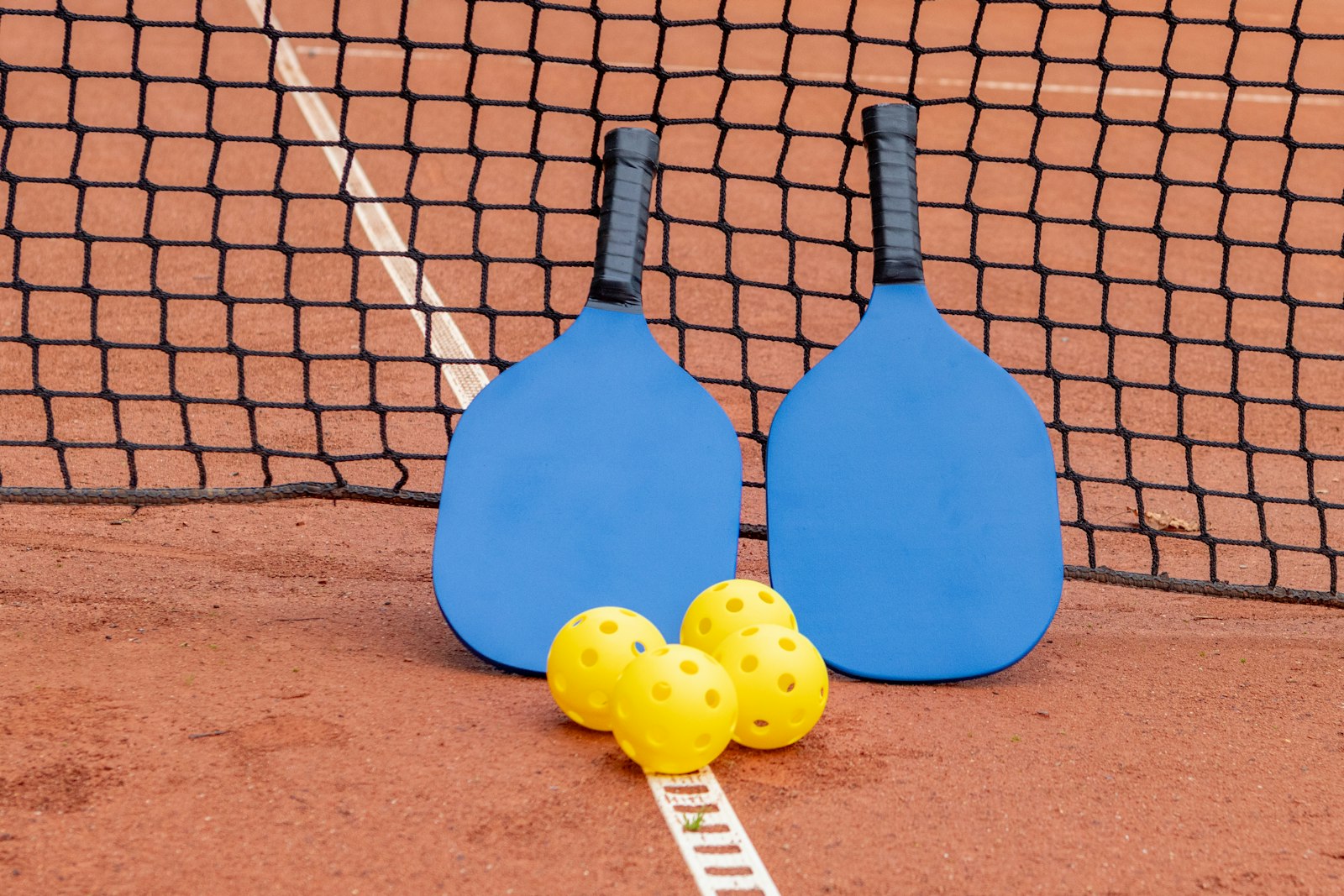Two paddles and a pair of yellow balls propped against the base of a net on an outdoor court.