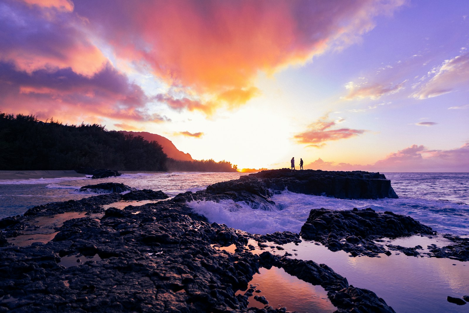 Two people watching a vivid sunset over rocks and surf on Kauai's north shore, one of the sunset spots on Kauai.