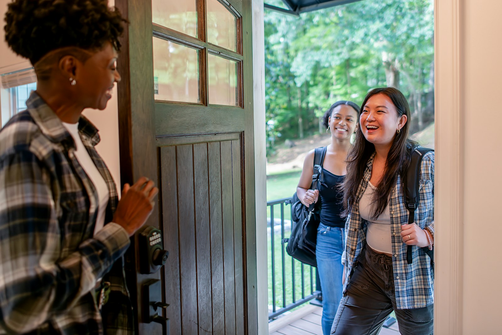 Someone opening a front door to two friends arriving with bags, all of them laughing.