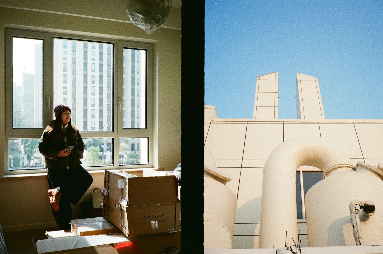 A person standing by a window beside unpacked moving boxes in a bare apartment, just moved in.