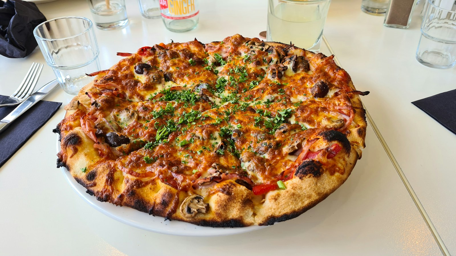 A pizza topped with herbs on a dining table beside glasses and cutlery.