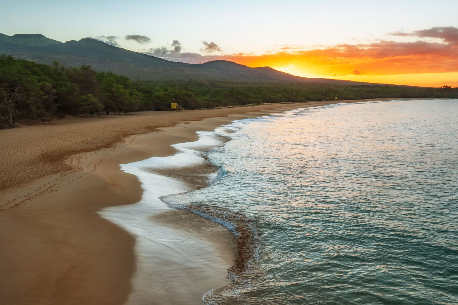 A quiet Maui beach at sunset, with gentle waves, forested hills, and a golden sky.