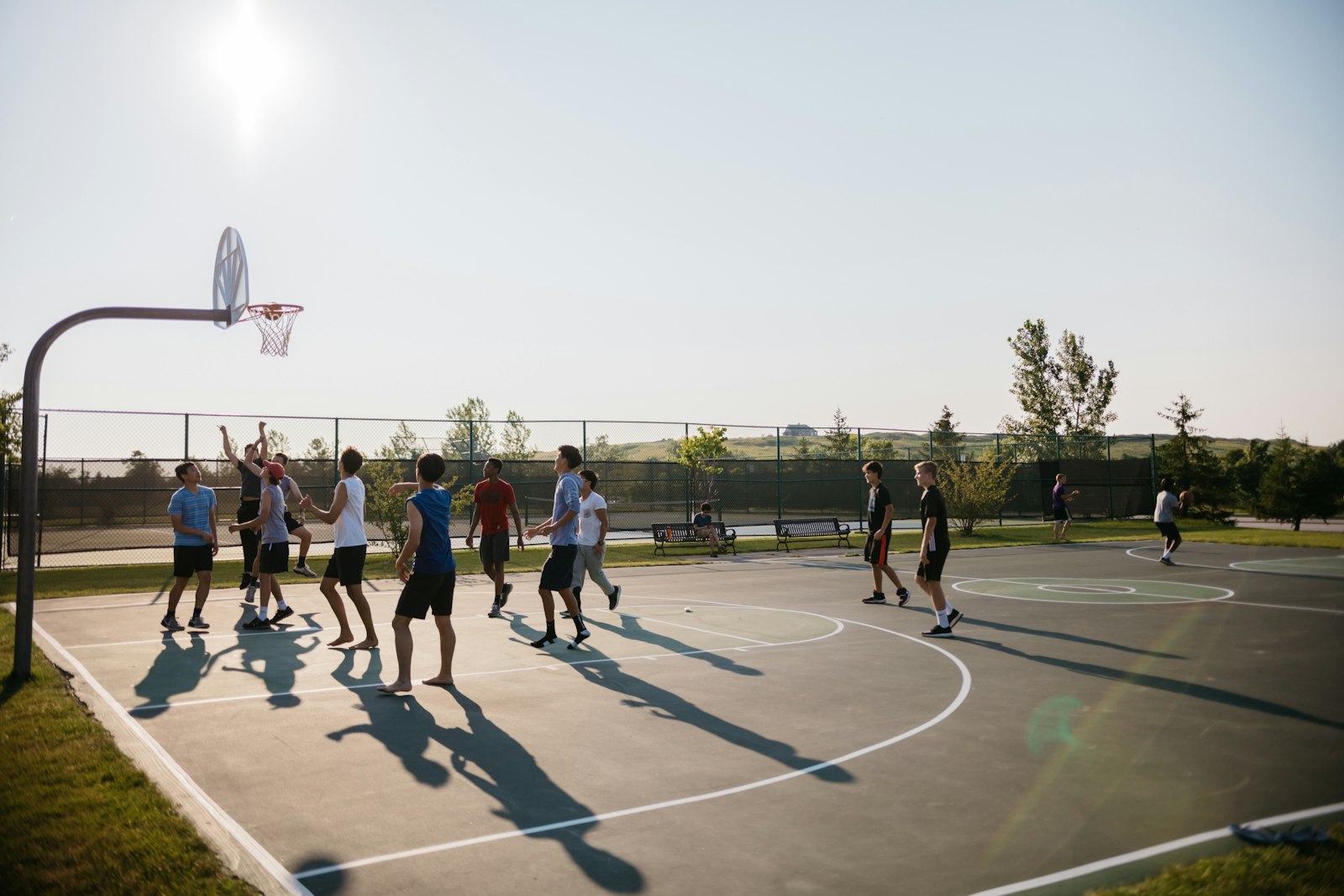 A pickup basketball game on an outdoor public court in late afternoon, players' shadows stretched long across the asphalt.