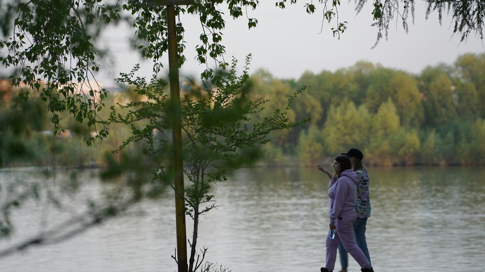 Two people walking beside water with leafy trees nearby.