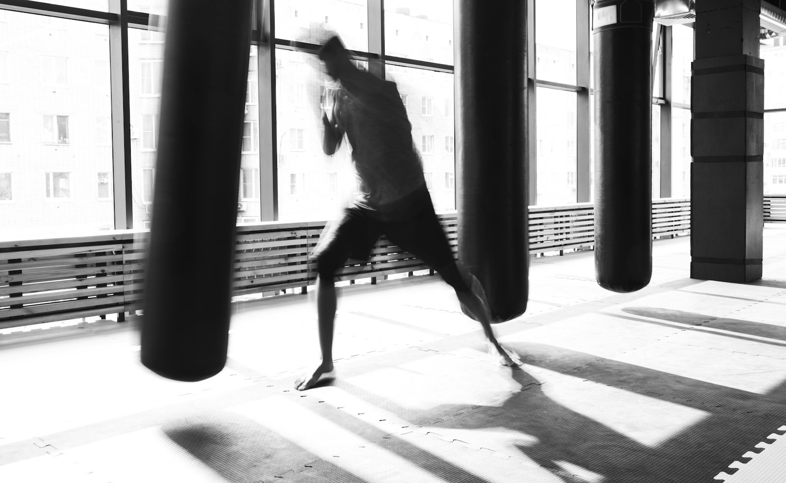 A lone figure working a heavy bag in a bright gym, silhouetted against tall windows.