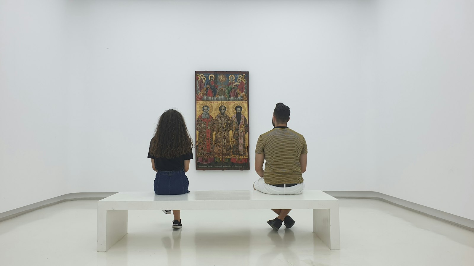 Two people sitting apart on the same gallery bench, both looking at one painting on a white wall.